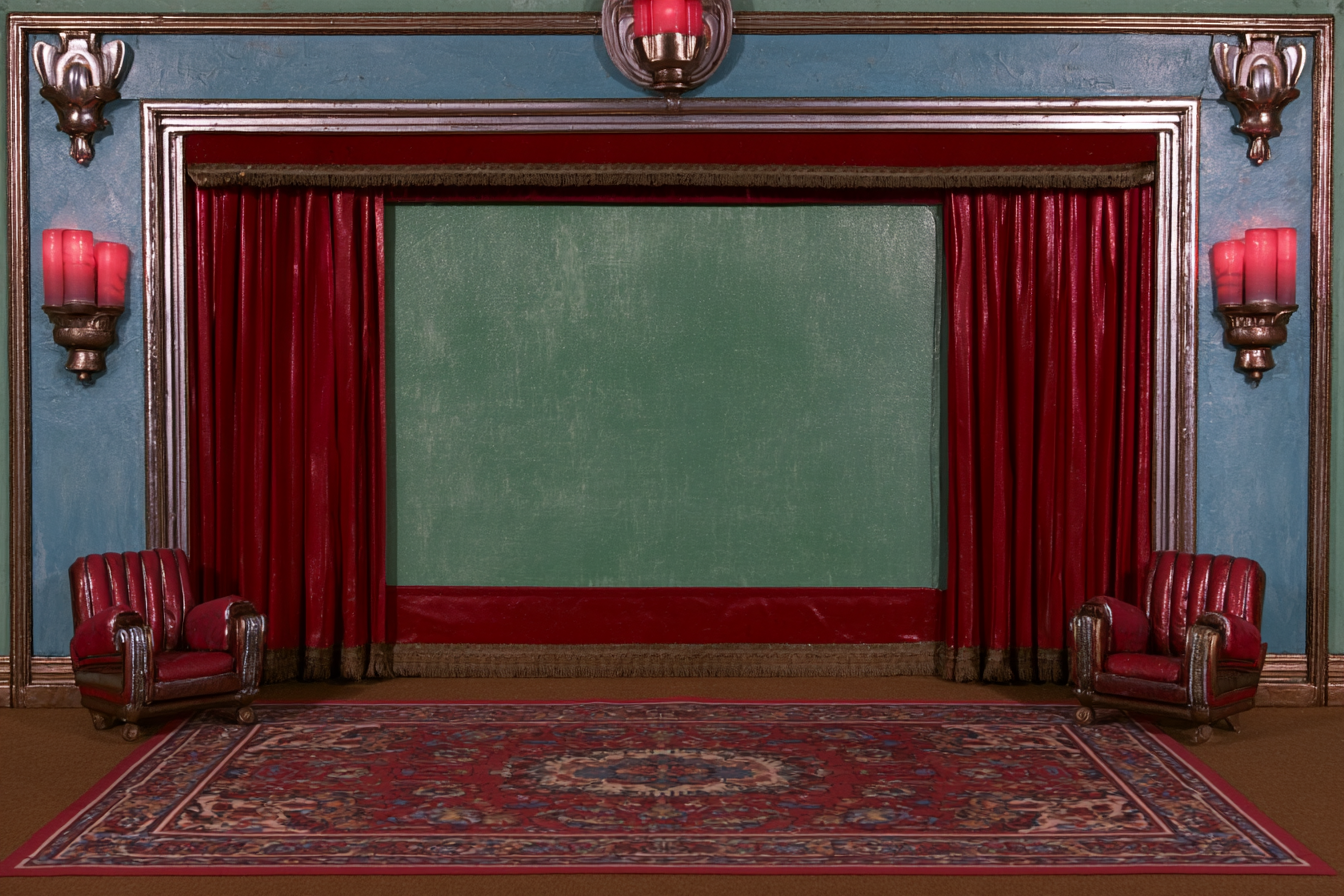An old cinema with red velvet curtains, candlelit walls, and two empty armchairs.